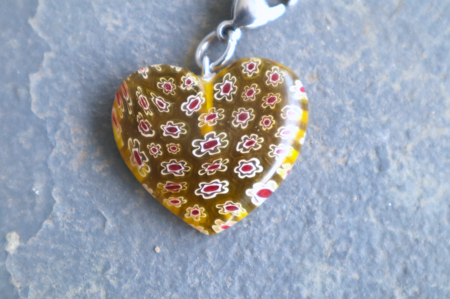A heart-shaped yellow pendant with white and red flower patterns. A silver clasp is attached to the top of the pendant, resting on a gray surface.