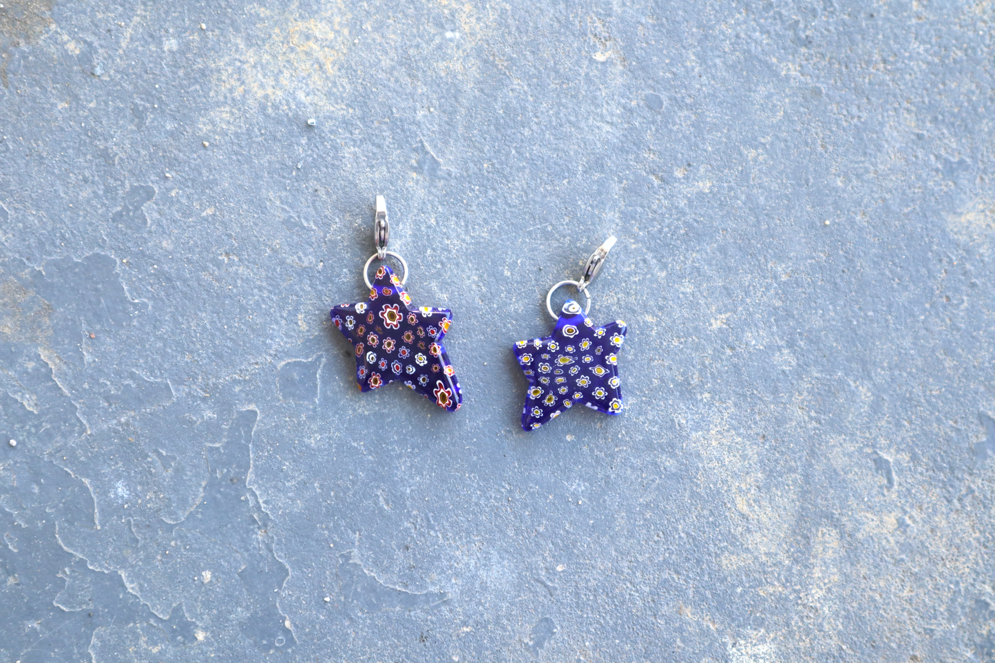 2 Star-shaped blue pendants with multi-colored flower patterns. A silver clasp is attached to the top of the pendant, resting on a gray surface.