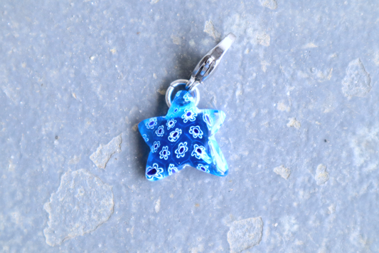 Star-shaped blue pendant with white and blue flower patterns. A silver clasp is attached to the top of the pendant, resting on a gray surface.