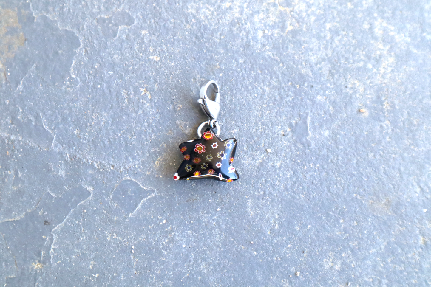 Star-shaped black pendant with white, red, yellow and blue flower patterns. A silver clasp is attached to the top of the pendant, resting on a gray surface.