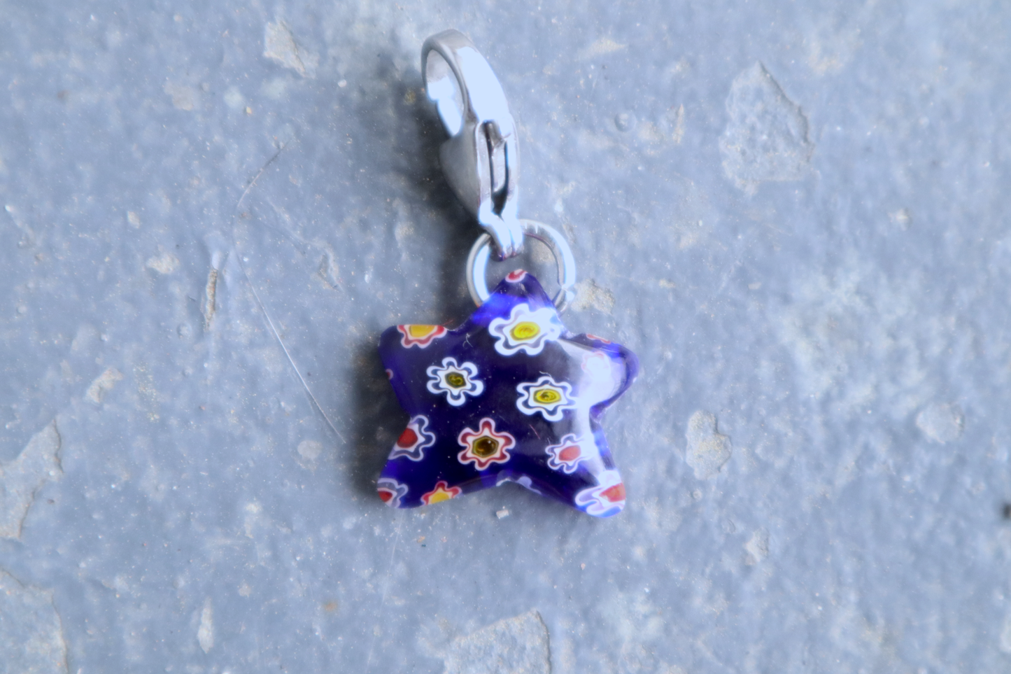 Star-shaped blue pendant with white, yellow and red flower patterns. A silver clasp is attached to the top of the pendant, resting on a gray surface.