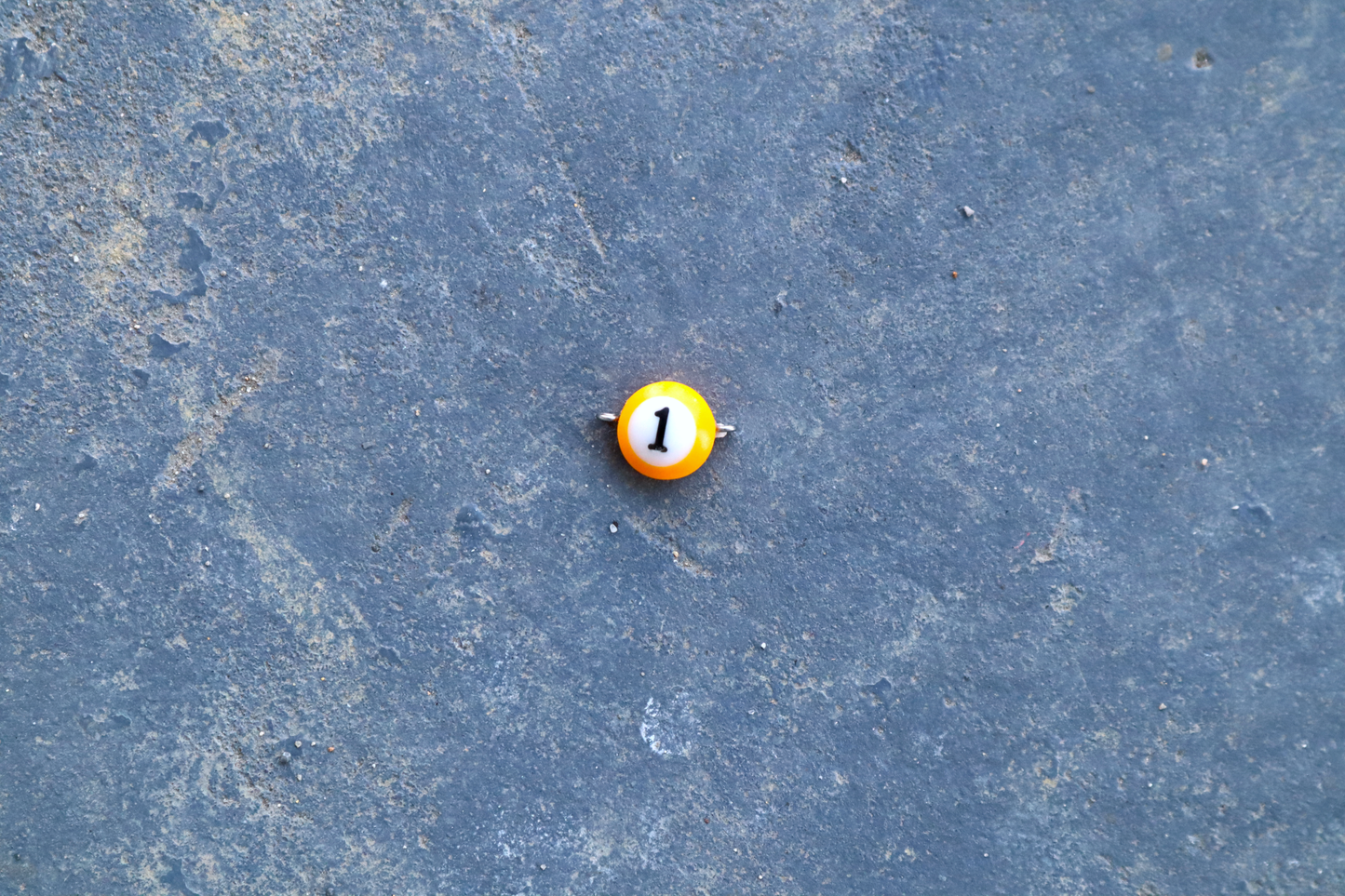 A yellow mini pool ball pendant with silver rings attached to the right and left side of the pendant A "1" is in the center of the pendant which is resting on a blue surface.