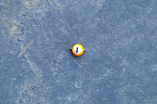 A yellow mini pool ball pendant with silver rings attached to the right and left side of the pendant A "1" is in the center of the pendant which is resting on a blue surface.