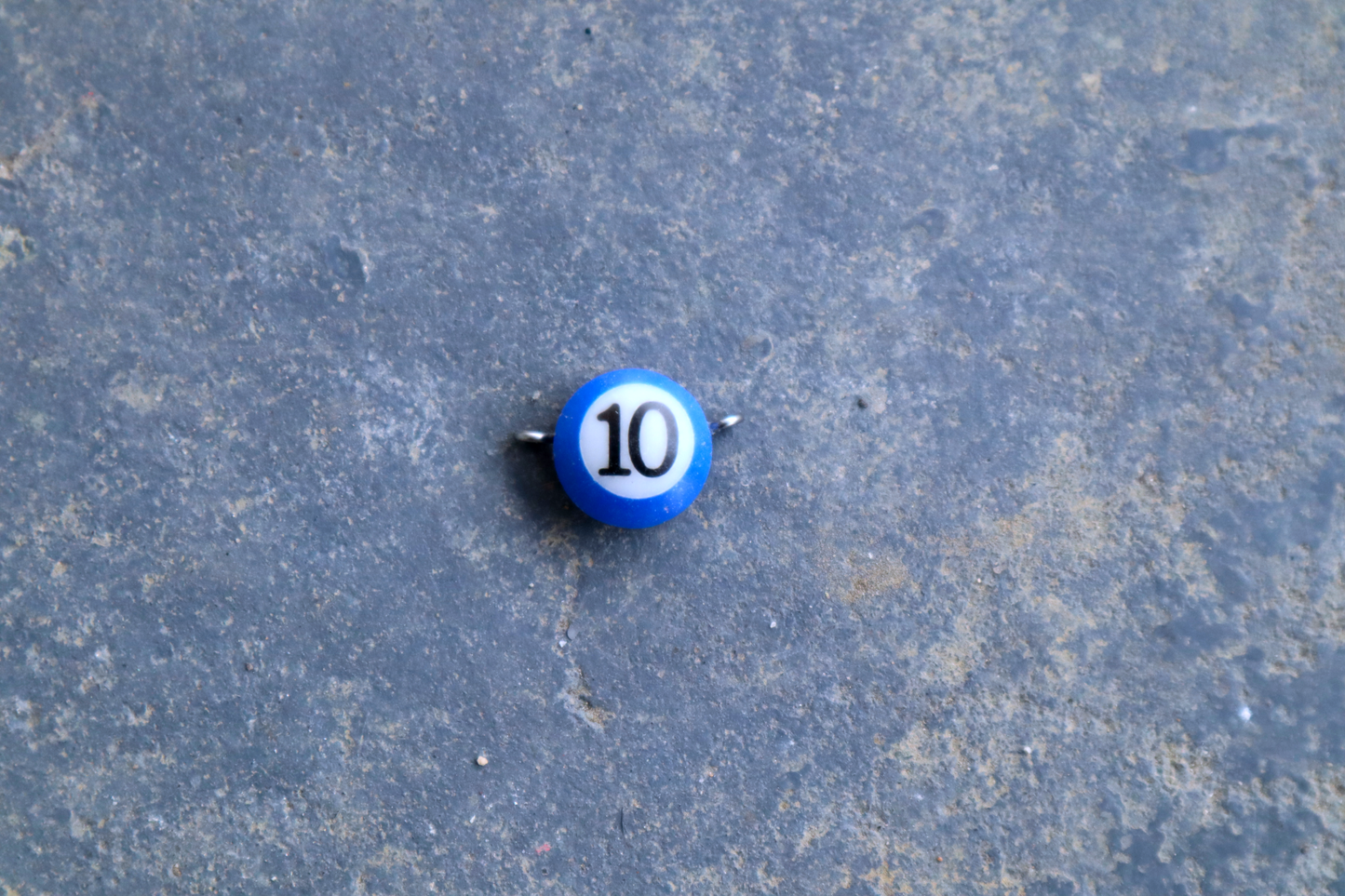 A blue mini pool ball pendant with silver rings attached to the right and left side of the pendant A "10" is in the center of the pendant which is resting on a blue surface.