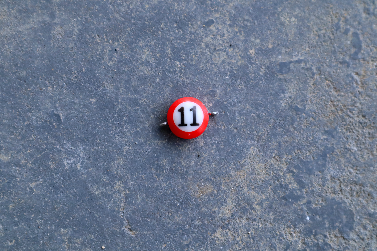 A red mini pool ball pendant with silver rings attached to the right and left side of the pendant A "11" is in the center of the pendant which is resting on a blue surface.