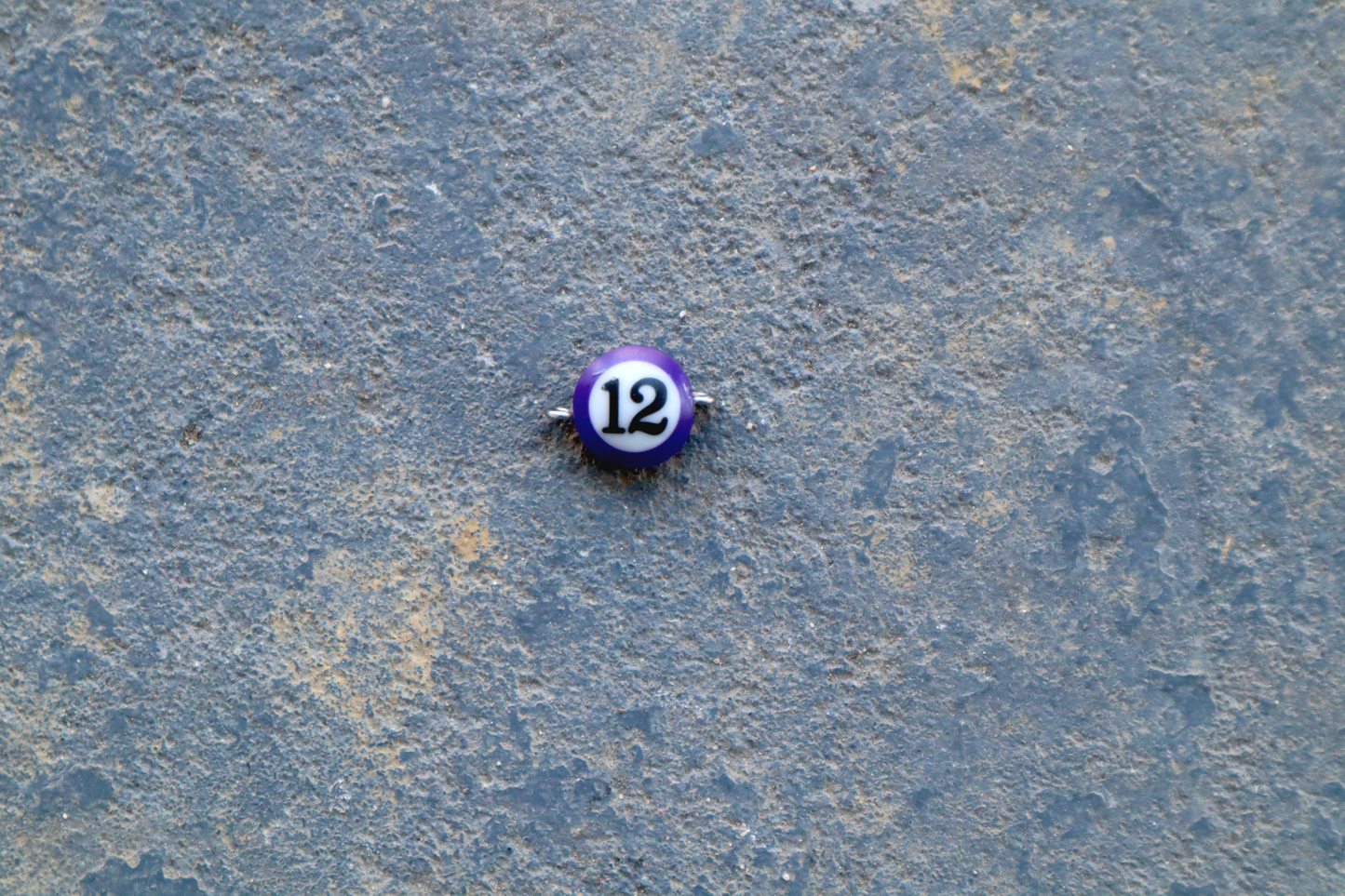 A purple mini pool ball pendant with silver rings attached to the right and left side of the pendant A "12" is in the center of the pendant which is resting on a blue surface.