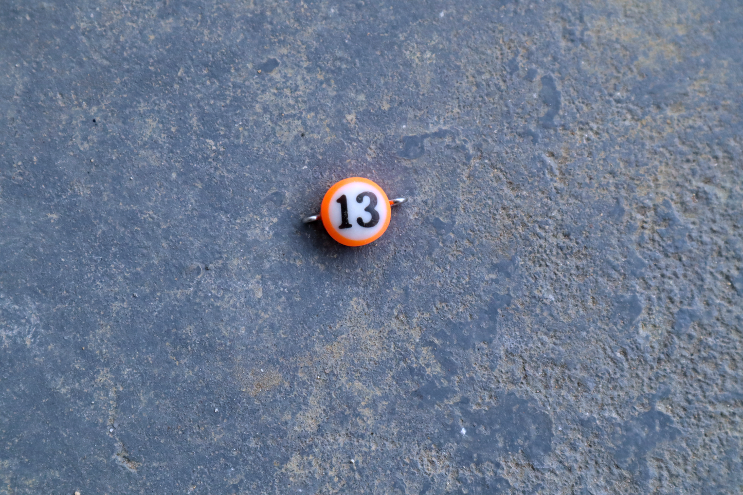 An orange mini pool ball pendant with silver rings attached to the right and left side of the pendant A "13" is in the center of the pendant which is resting on a blue surface.