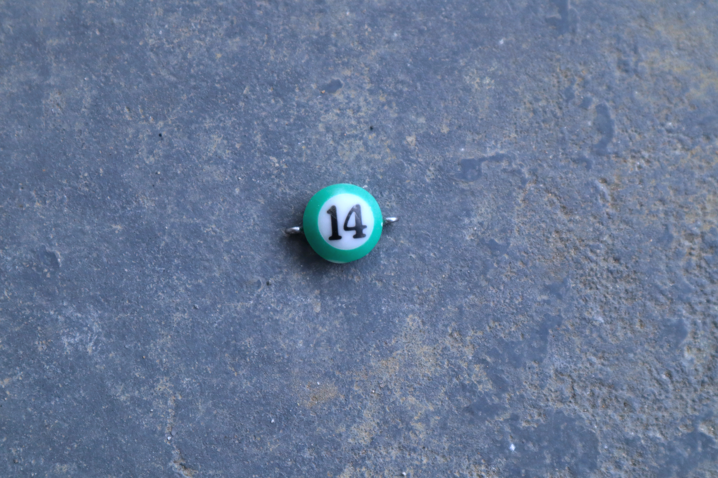 A green mini pool ball pendant with silver rings attached to the right and left side of the pendant A "14" is in the center of the pendant which is resting on a blue surface.