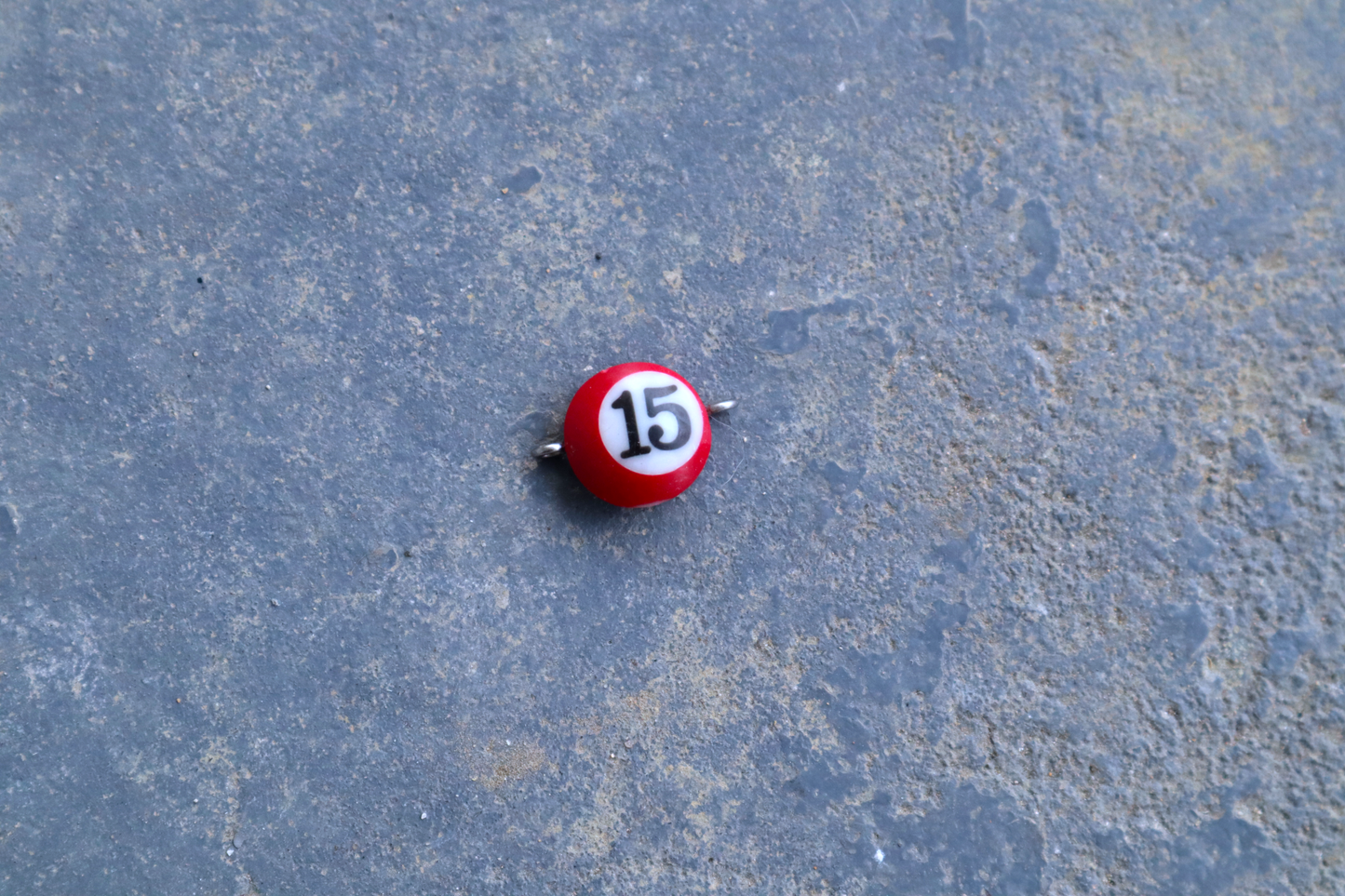 A red mini pool ball pendant with silver rings attached to the right and left side of the pendant A "15" is in the center of the pendant which is resting on a blue surface.