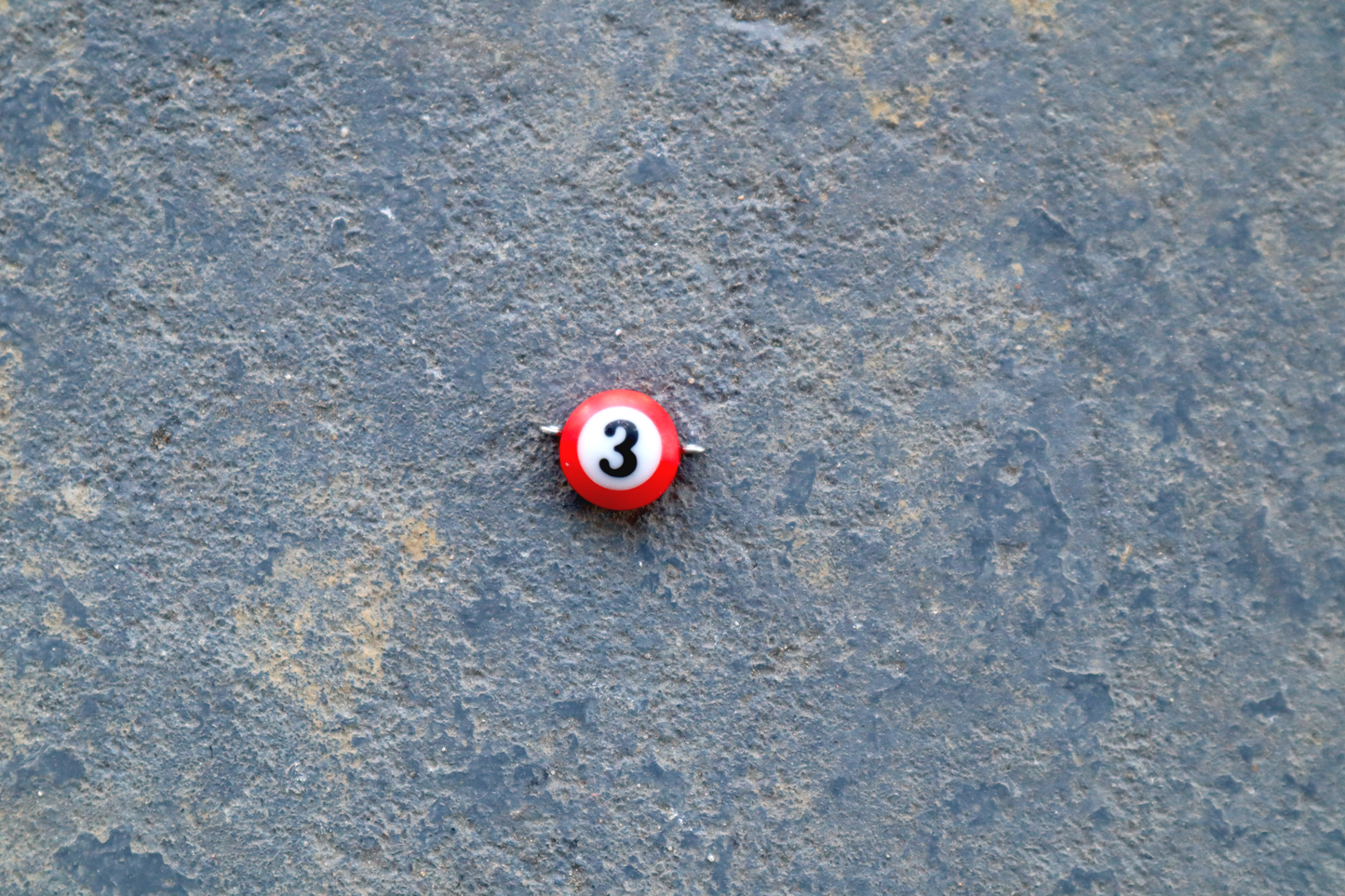 A red mini pool ball pendant with silver rings attached to the right and left side of the pendant A "3" is in the center of the pendant which is resting on a blue surface.