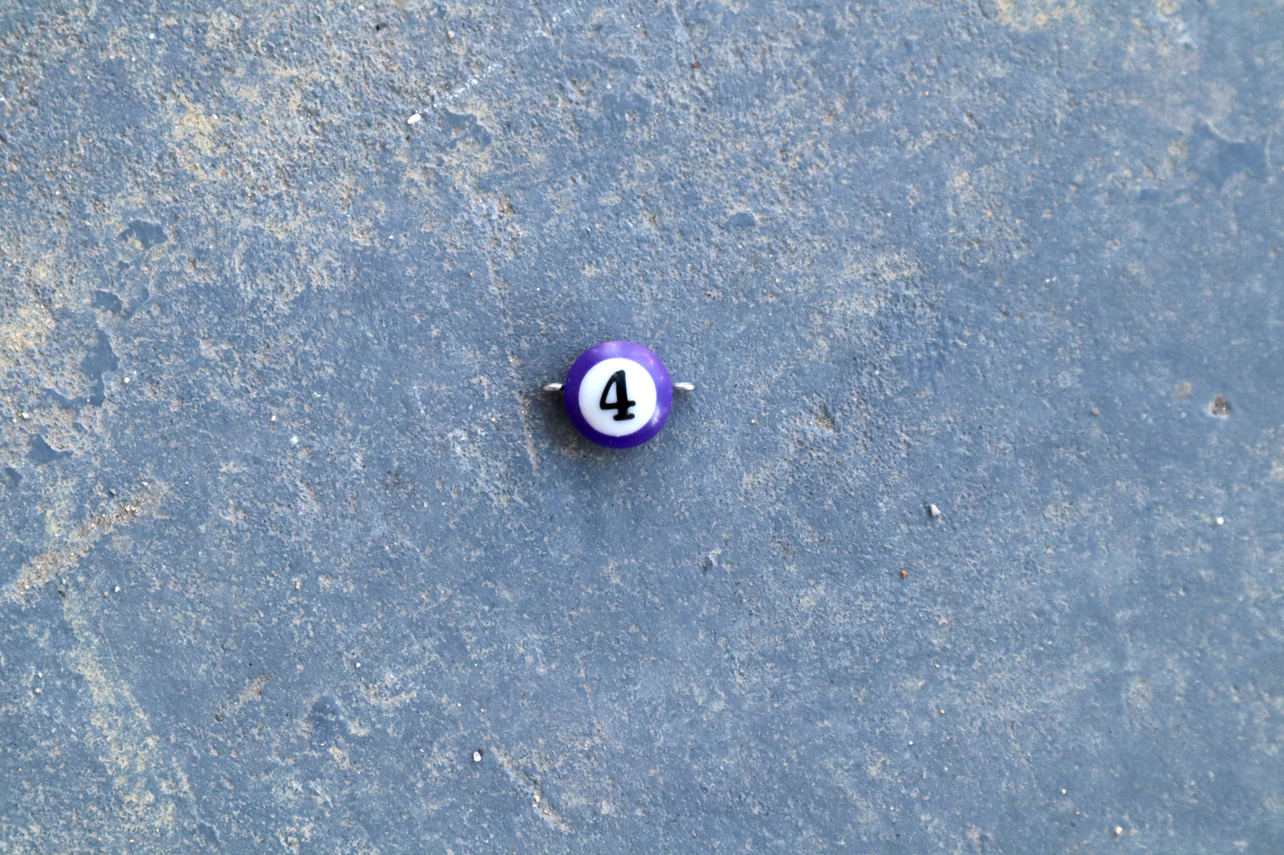 A purple mini pool ball pendant with silver rings attached to the right and left side of the pendant A "4" is in the center of the pendant which is resting on a blue surface.