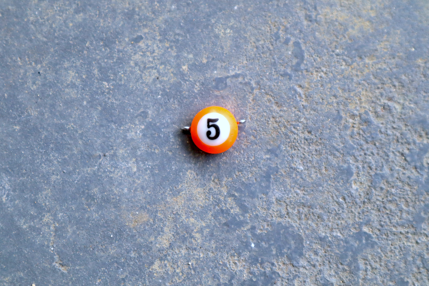 An orange mini pool ball pendant with silver rings attached to the right and left side of the pendant A "5" is in the center of the pendant which is resting on a blue surface.