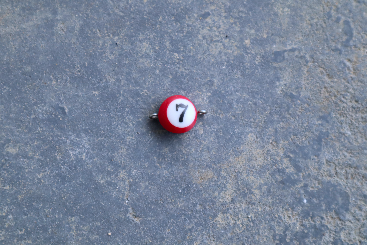 A red mini pool ball pendant with silver rings attached to the right and left side of the pendant A "7" is in the center of the pendant which is resting on a blue surface.