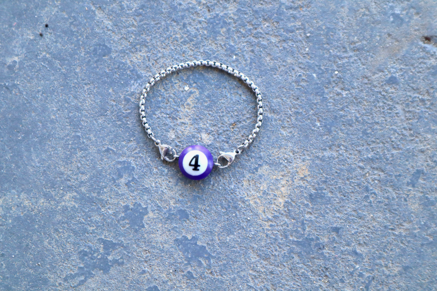 Silver chain bracelet with a purple billiard ball pendant marked "4" laid against a blue/gray surface.