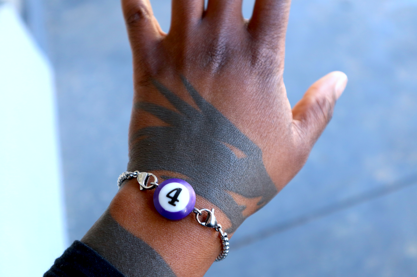 A hand with a dragon tattoo wears a bracelet with a purple mini pool ball charm numbered 4