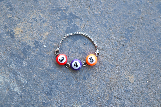 Silver chain bracelet with a red, purple, and orange billiard ball pendant marked "3", "4", and "5" laid against a blue/gray surface.