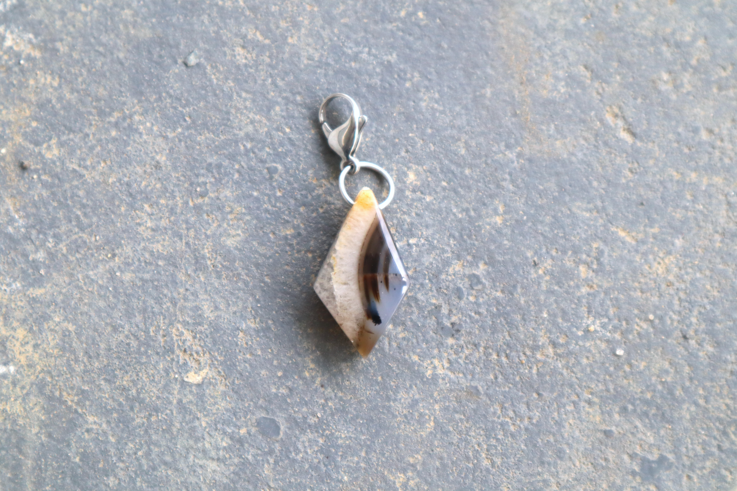 A clear, white and blue diamond-shaped stone with black streaks running down the right side of the stone. A silver clasp is attached to the top of the pendant, resting on a gray surface.