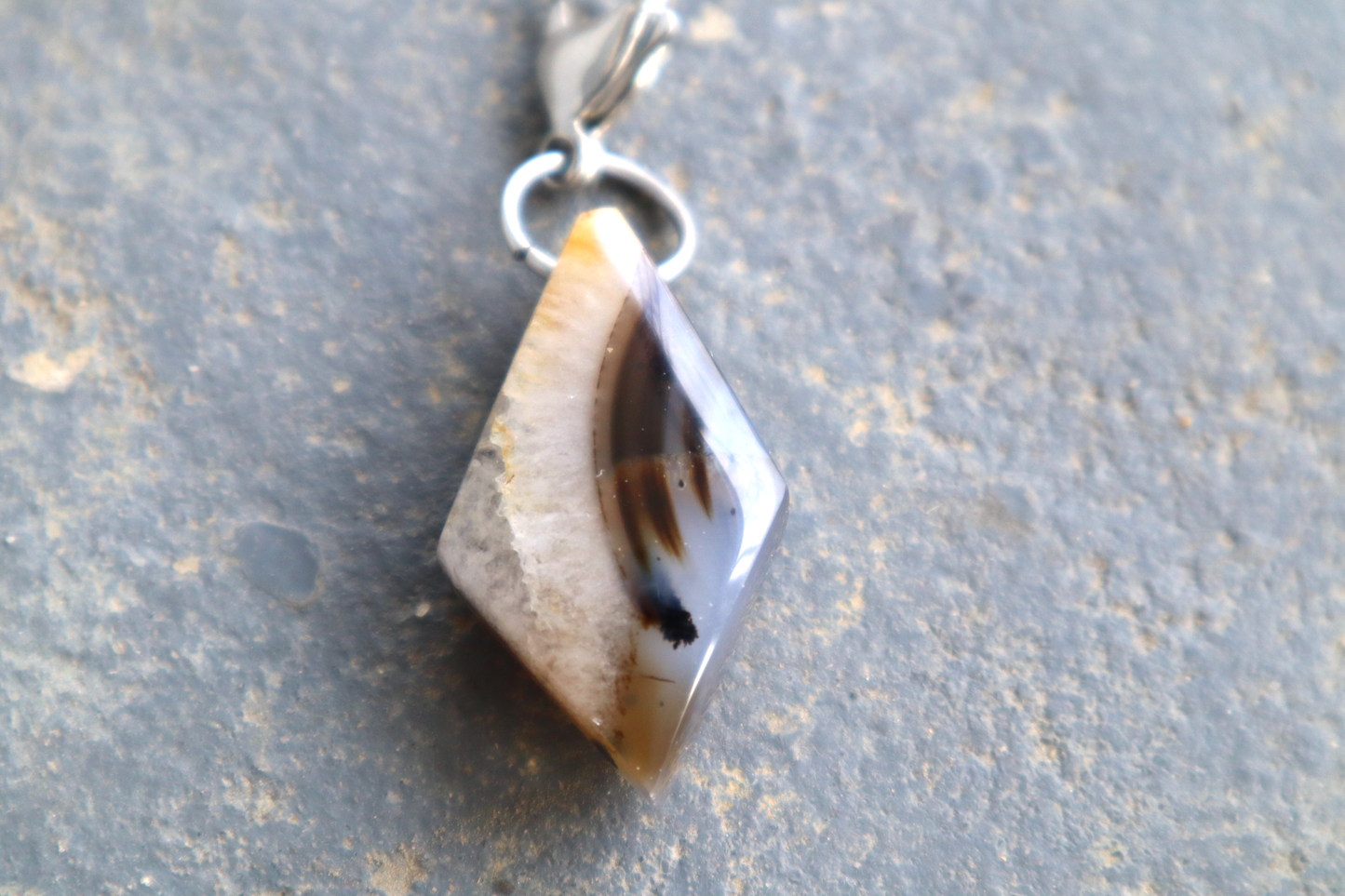 A clear, white and blue diamond-shaped stone with black streaks running down the right side of the stone. A silver clasp is attached to the top of the pendant, resting on a gray surface.