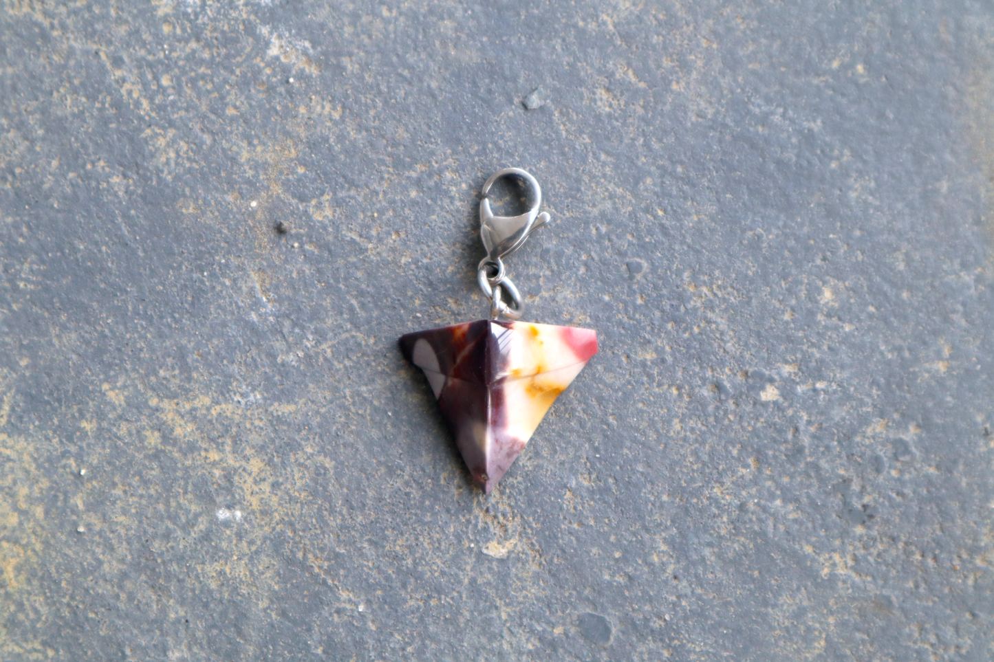 purple, white, yellow and tan triangular shaped stone with a silver clasp attached to the top of the pendant, resting on a blue/gray surface.