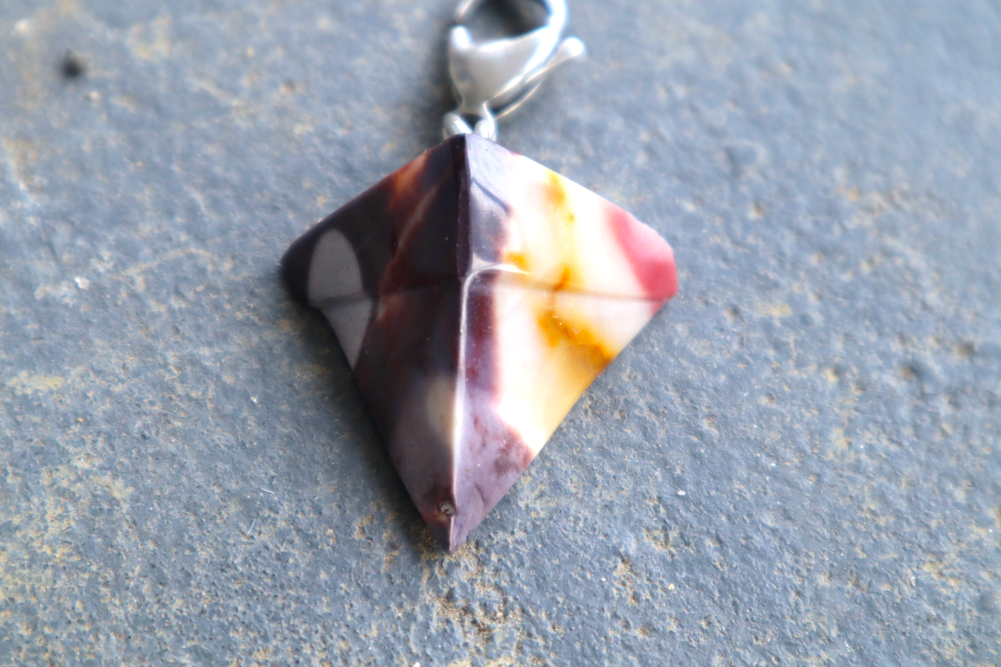 purple, white, yellow and tan triangular shaped stone with a silver clasp attached to the top of the pendant, resting on a blue/gray surface.