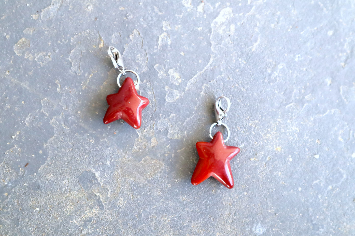 two five-pointed star shaped stones with stainless-steel clasps attached, against a blue/grey stone background. The stone pendants are a scarlet to bright cherry red color. The pendant on the left is shorter than the elongated pendant on the right.