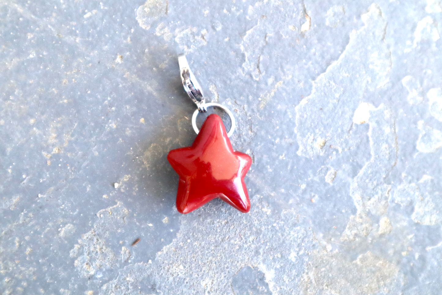 A five-pointed star shaped stone with a stainless-steel clasp attached to it, against a blue/grey stone background. The stone pendant is a scarlet to bright cherry red color. the middle right point on this star is shorter than the other four points. The bottom right point is thinner than the other fur points.