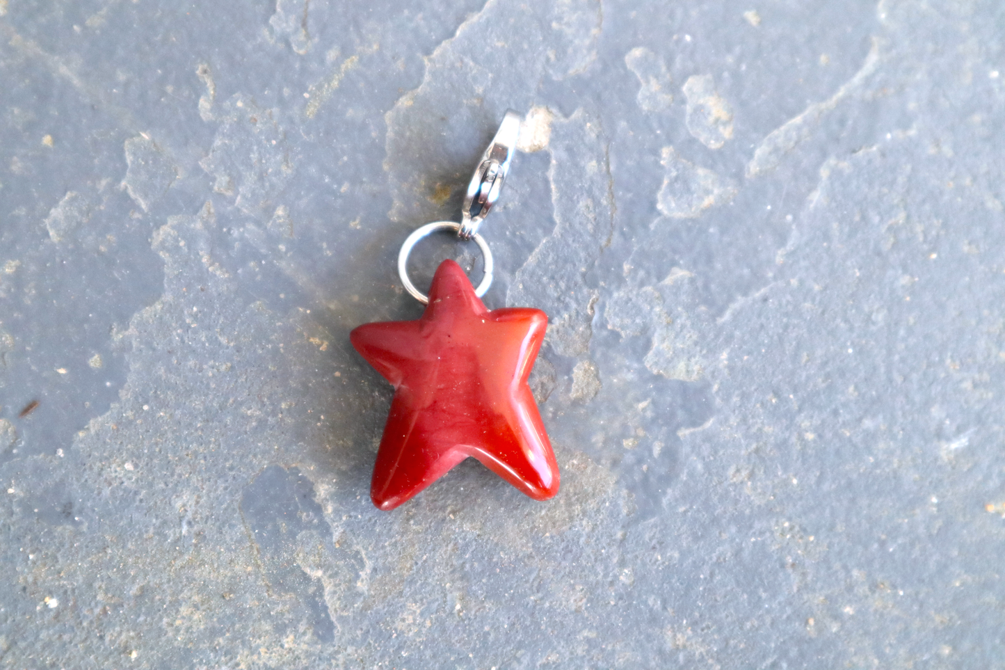 A five-pointed star shaped stone with a stainless-steel clasp attached to it, against a blue/grey stone background. The stone pendant is a scarlet to bright cherry red color with a stripe of pink running down the middle. The two bottom points are longer than the other three points.