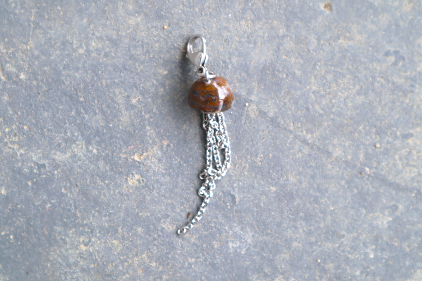 A mushroom cap-shaped stone with steel chains hanging from the bottom of it and a steel clasp attached to the top, against a blue/grey stone background. The stone pendant is orange and red.