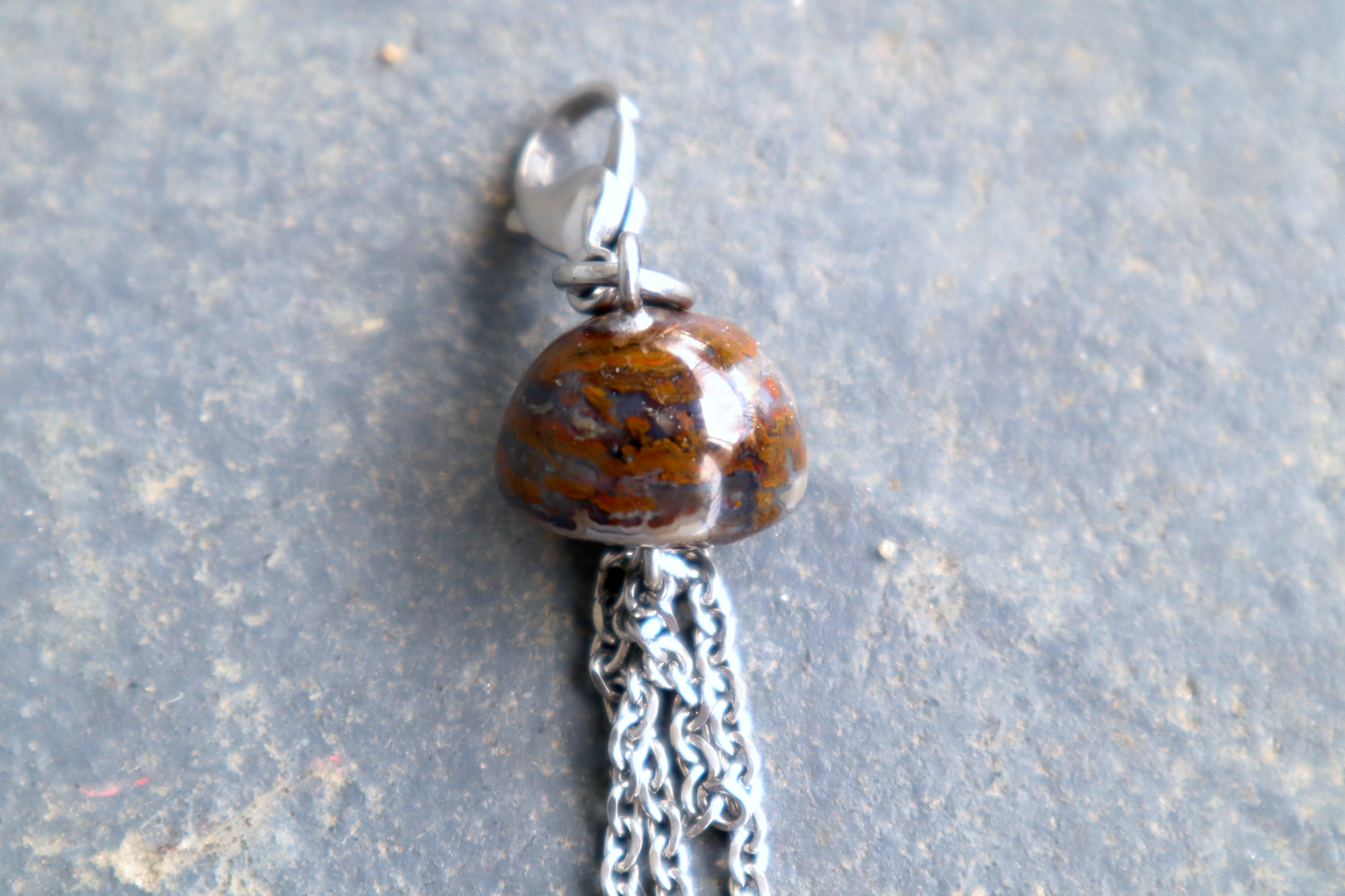 A mushroom cap-shaped stone with steel chains hanging from the bottom of it and a steel clasp attached to the top, against a blue/grey stone background. The stone pendant is orange and red.