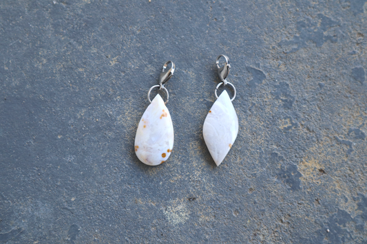 a pointed oval-shaped stone and a tear drop-shaped stone, both with a stainless-steel clasp attached to it, against a blue/grey stone background. The stone pendants are light grey, light blue, and white in color, with brown dots scattered across their surface.