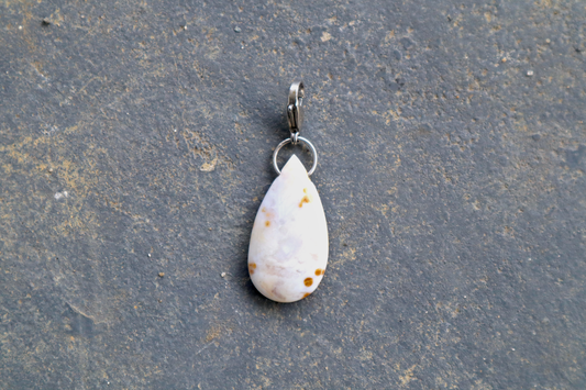 a tear drop-shaped stone, with a stainless-steel clasp attached to it, against a blue/grey stone background. The stone pendant is light grey, light blue, and white in color, with brown dots scattered across its surface.
