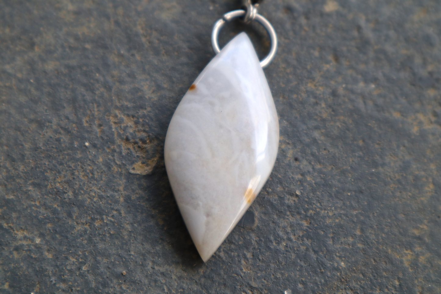 a close-up picture of a pointed oval-shaped stone, with a stainless-steel clasp attached to it, against a blue/grey stone background. The stone pendant is light grey, light blue, and white in color, with 2 brown dots on its surface.