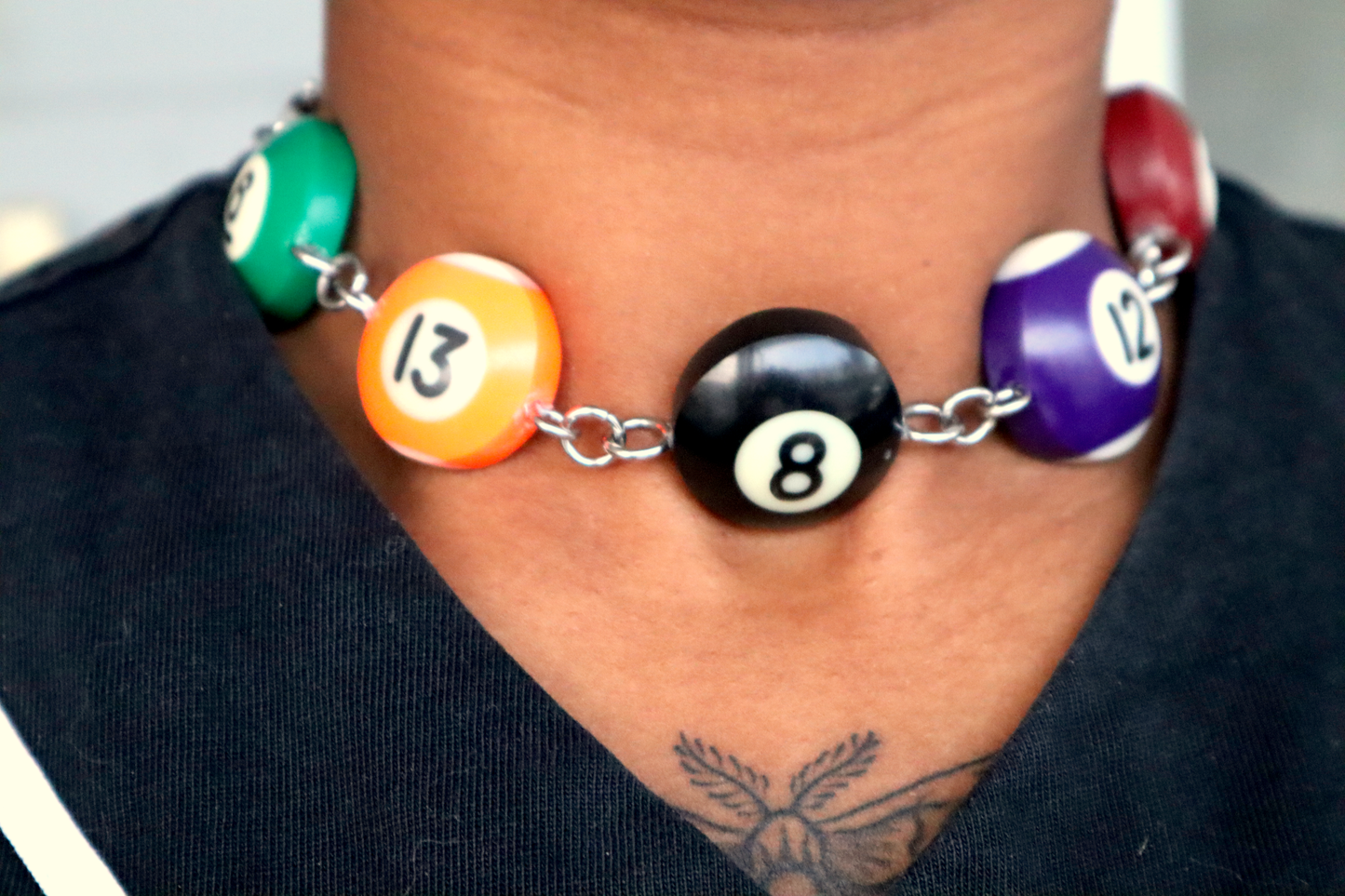A choker made of pool ball charms numbered 6, 13, 8, 12 and 7 being worn on a neck. The choker has a steel chain and hardware to connect the charms.