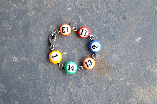 A bracelet of mini pool ball charms numbered 1, 14, 13, 2, 11 and 13. The bracelet has steel hardware to connect the charms.