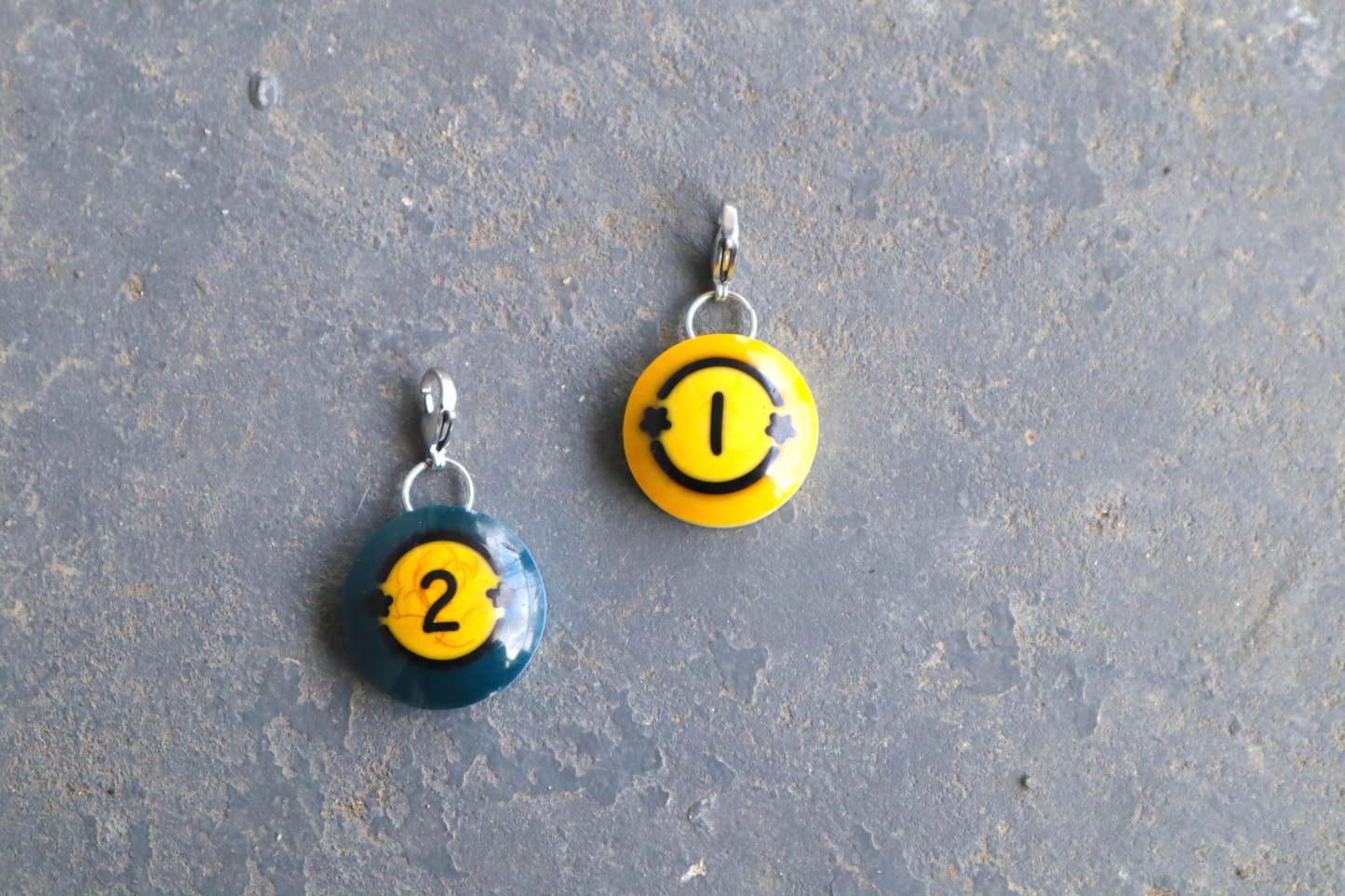 Two billiard ball pendants marked "1" and "2" with 2 black stars on both sides of the numbers. A silver clasp is attached to the tops of the pendants against a blue/gray surface