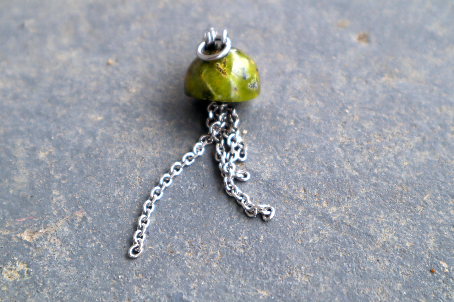 A mushroom cap-shaped stone with steel chains hanging from the bottom of it and a steel clasp attached to the top, against a blue/grey stone background. The stone pendant is green.