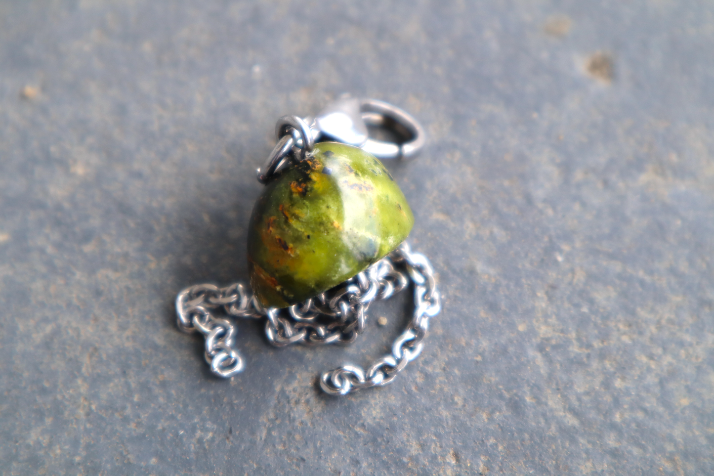 A mushroom cap-shaped stone with steel chains hanging from the bottom of it and a steel clasp attached to the top, against a blue/grey stone background. The stone pendant is orange and green.