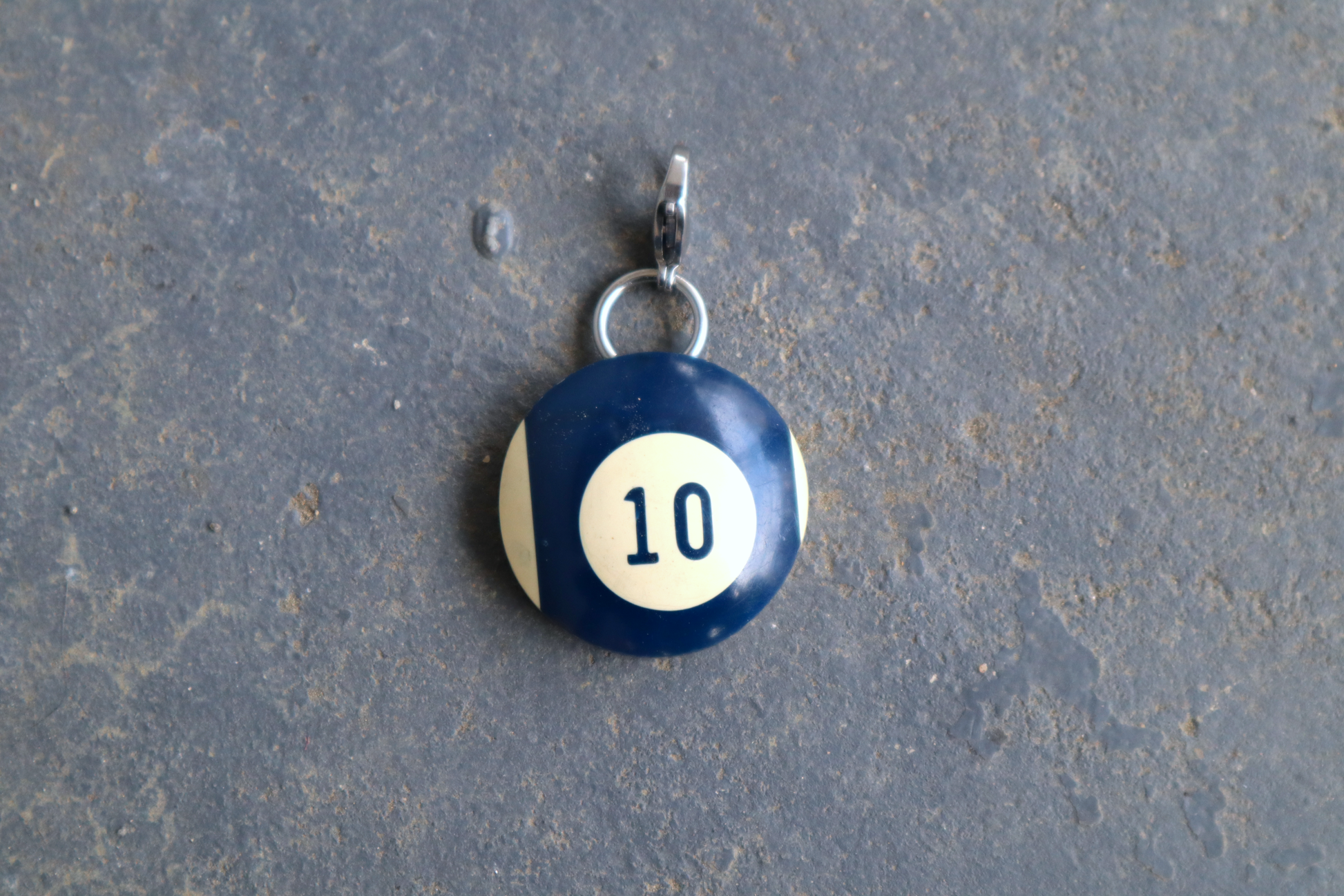 a blue and white billiard ball pendant marked "10" in the center. A silver clasp is attached to the top of the pendant against a blue/gray surface