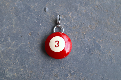 a red billiard ball pendant marked "3" in the center. A silver clasp is attached to the top of the pendant against a blue/gray surface