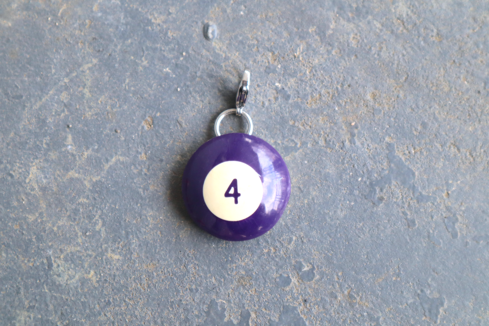 a purple billiard ball pendant marked "4" in the center. A silver clasp is attached to the top of the pendant against a blue/gray surface