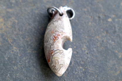grey, white, black and red a pointed oval-shaped stone with a silver clasp attached to the top of the pendant, resting on a gray surface.