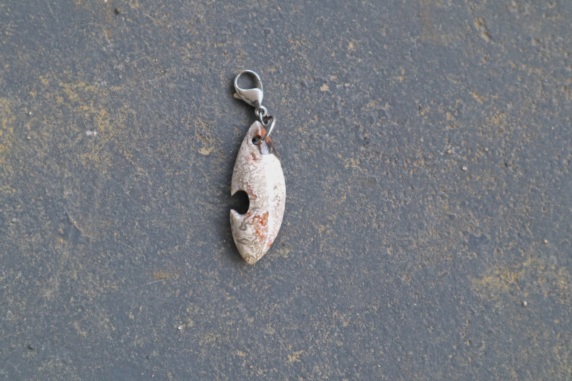 grey, white, black and red a pointed oval-shaped stone with a silver clasp attached to the top of the pendant, resting on a gray surface.