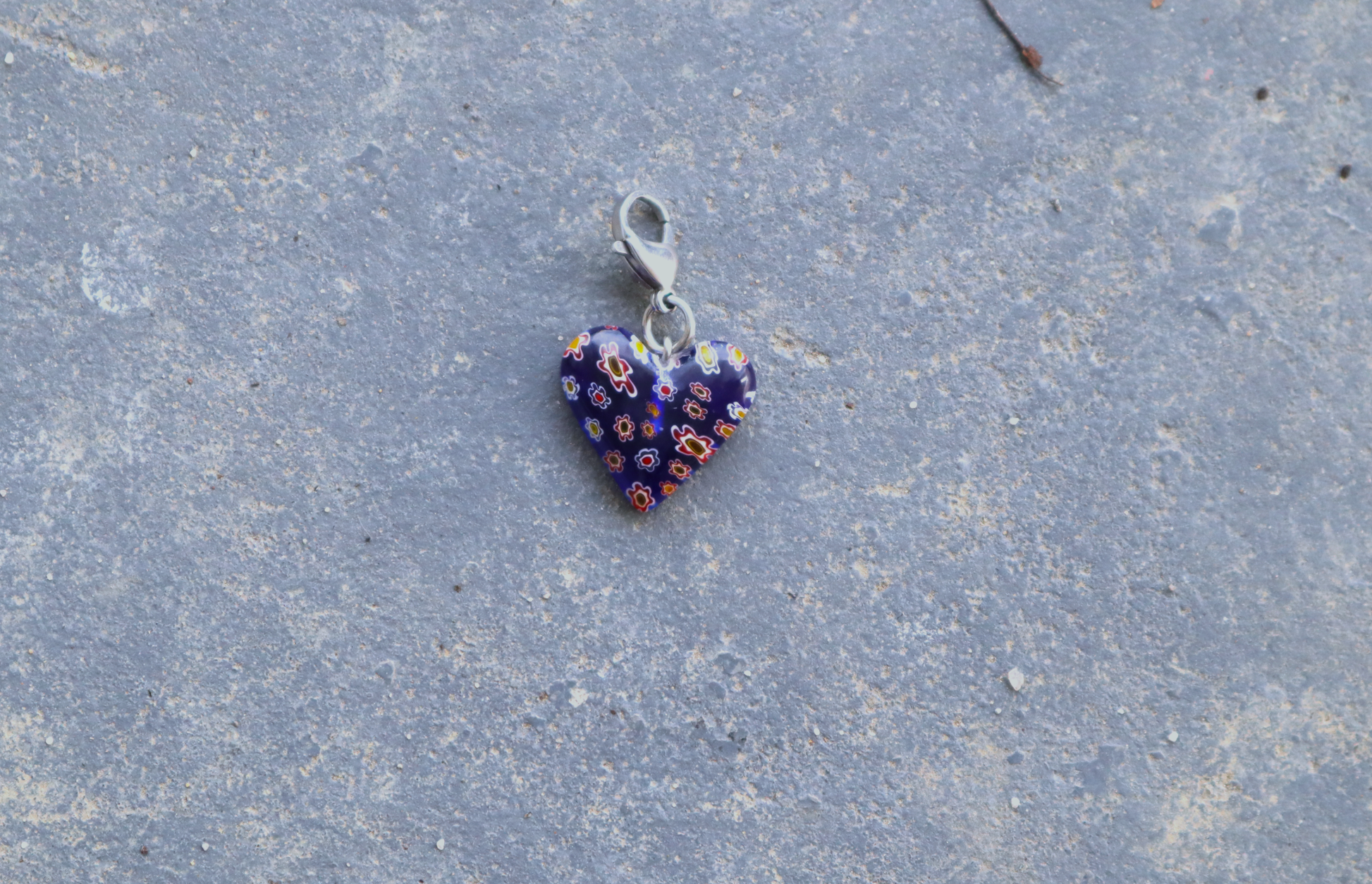 A heart-shaped blue pendant with white, red and yellow flower patterns. A silver clasp is attached to the top of the pendant, resting on a gray surface.