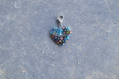 A heart-shaped multi-colored pendant with multi-colored flower patterns. A silver clasp is attached to the top of the pendant, resting on a gray surface.