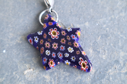 Star-shaped blue pendant with white, yellow and red flower patterns. A silver clasp is attached to the top of the pendant, resting on a gray surface.
