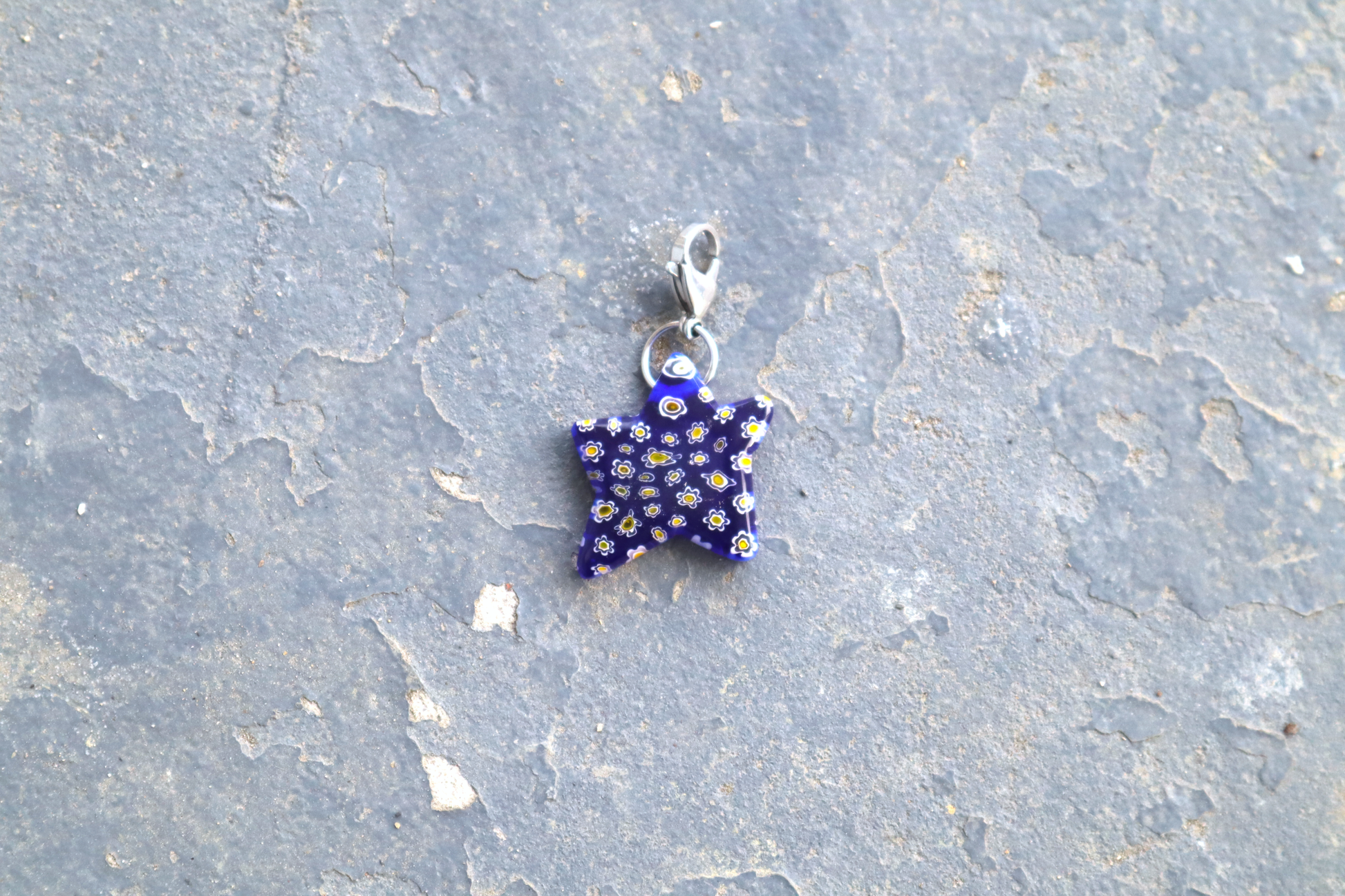 Star-shaped blue pendant with white and yellow flower patterns. A silver clasp is attached to the top of the pendant, resting on a gray surface.