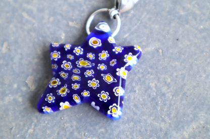 Star-shaped blue pendant with white and yellow flower patterns. A silver clasp is attached to the top of the pendant, resting on a gray surface.
