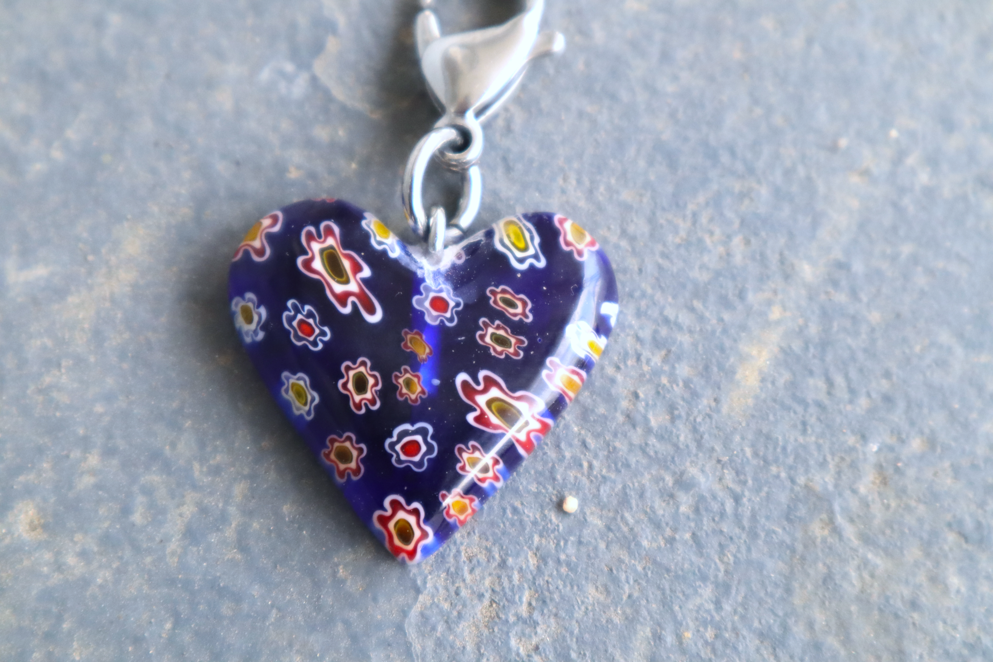 A heart-shaped blue pendant with white, red and yellow flower patterns. A silver clasp is attached to the top of the pendant, resting on a gray surface.