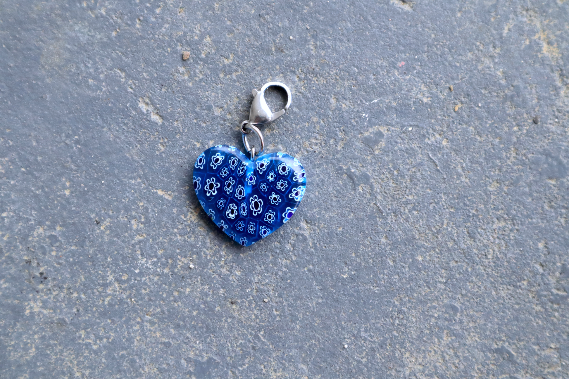 A heart-shaped light blue pendant with white and blue flower patterns. A silver clasp is attached to the top of the pendant, resting on a gray surface.