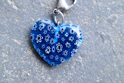 A heart-shaped light blue pendant with white and blue flower patterns. A silver clasp is attached to the top of the pendant, resting on a gray surface.